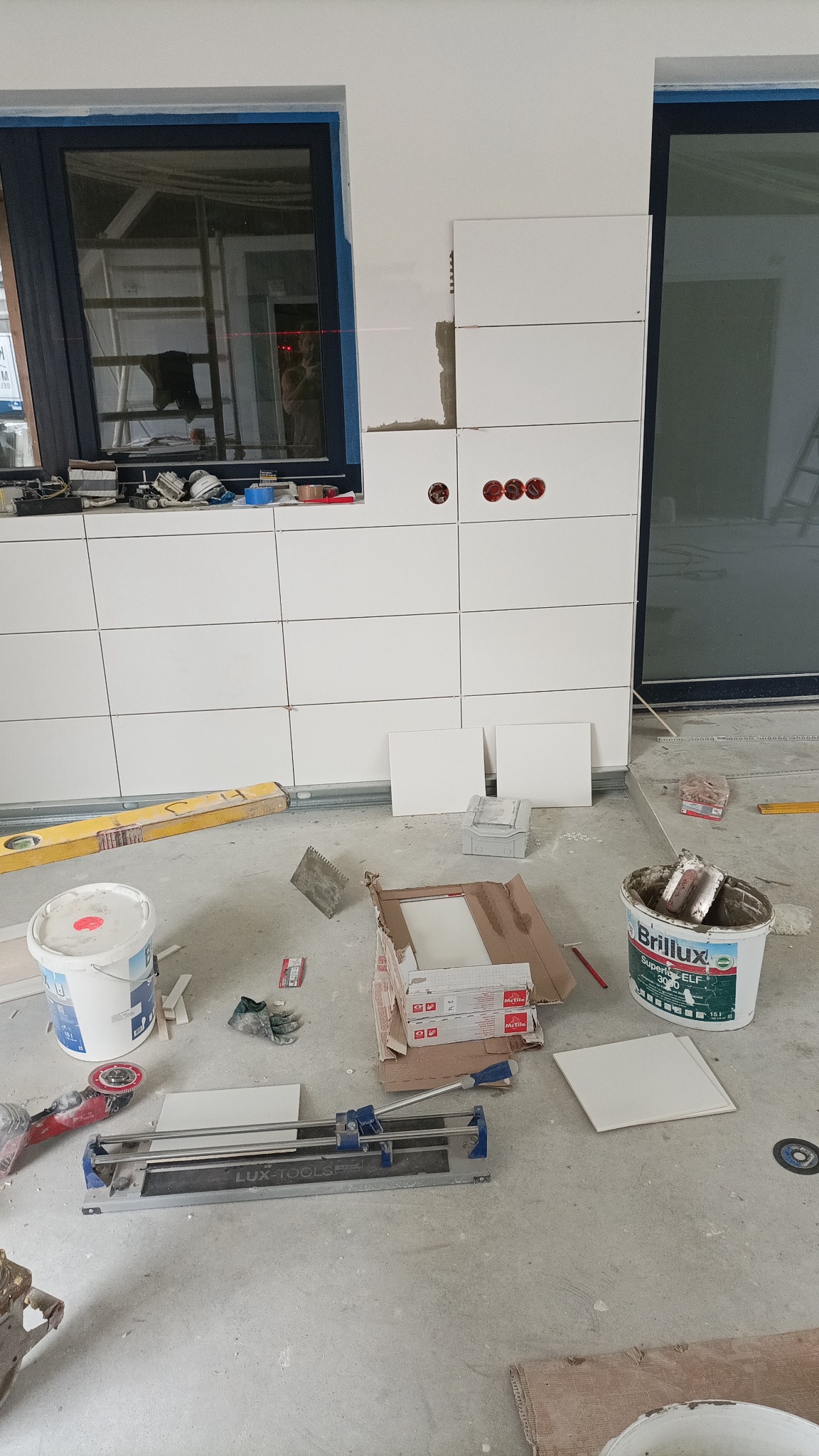 Świeżo ułożone białe płytki na ścianie, widoczne puszki elektryczne, poziomica, wiadro z klejem Brillux, przecinarka do płytek Lux Tools i kartony z płytkami, remont w toku.