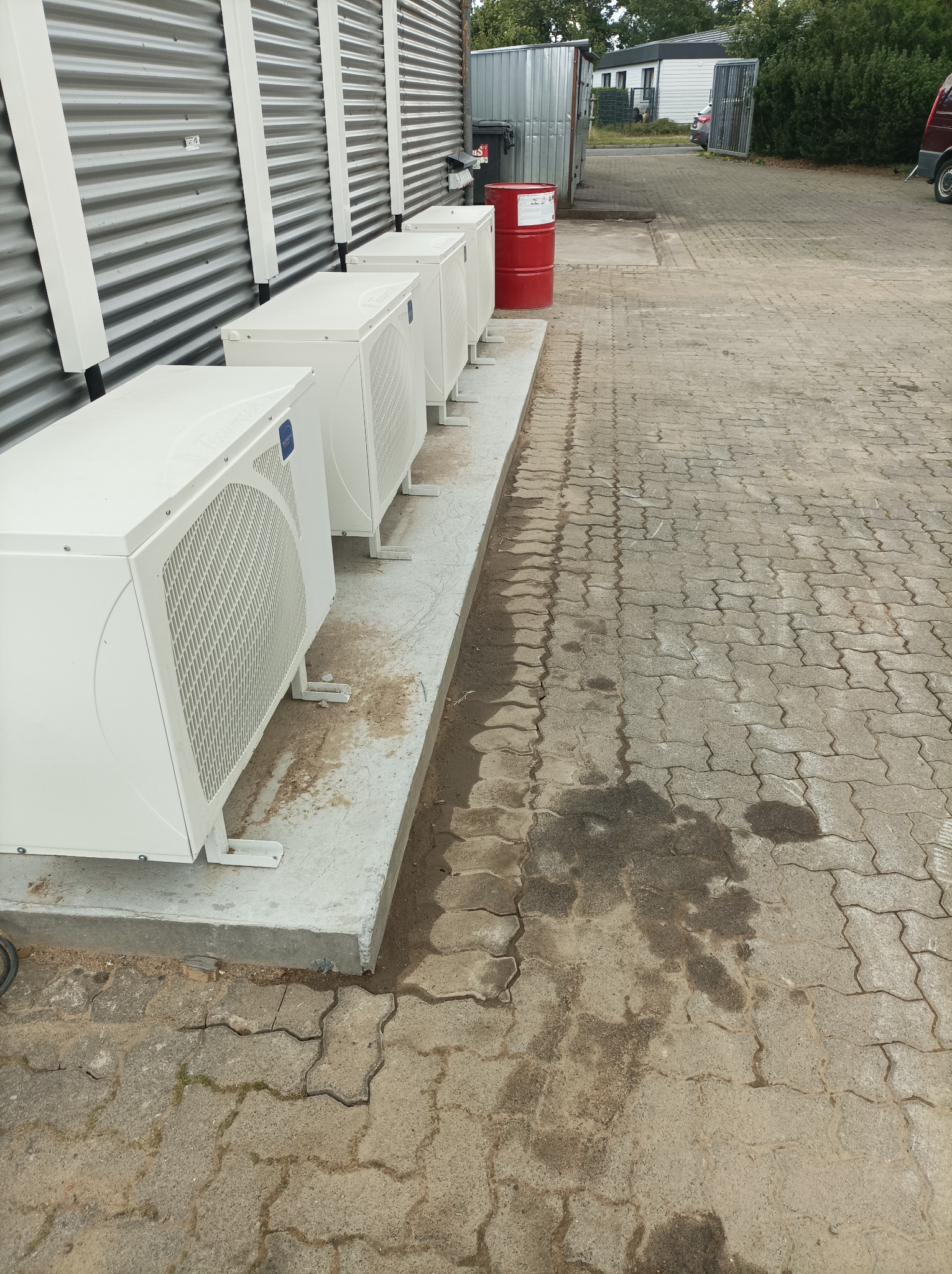 Rząd białych klimatyzatorów przemysłowych, ustawionych na betonowej podstawie wzdłuż blaszanej ściany, z czerwoną beczką i kostką brukową w tle.