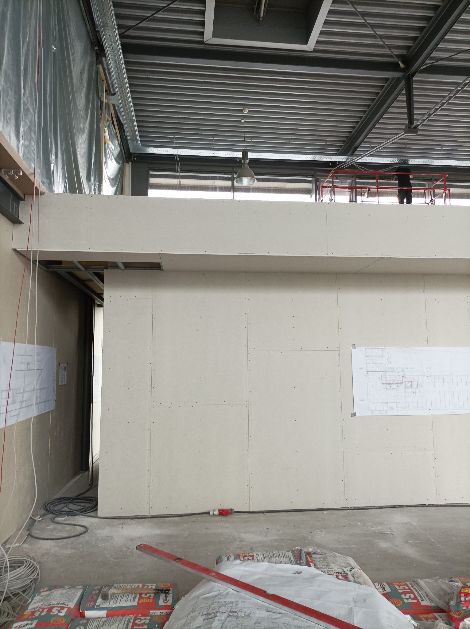 Wnętrze w trakcie remontu: ściany z płyt gipsowo-kartonowych, widoczne szpachlowane łączenia, plany architektoniczne na ścianie, worki zaprawy na podłodze i pracownik na podnośniku w tle.
