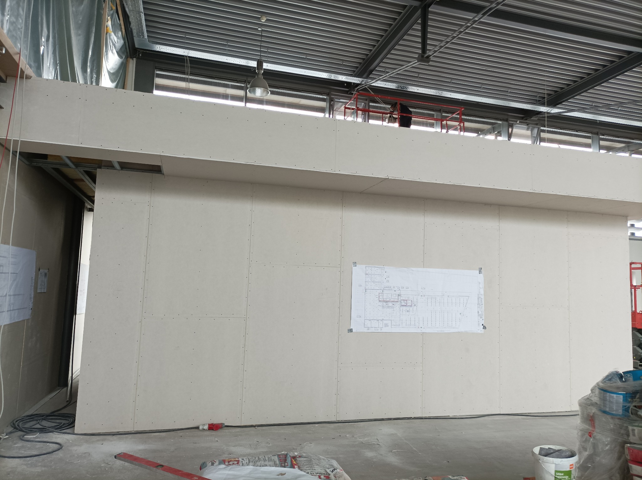 Wnętrze hali w trakcie prac wykończeniowych: ściany z płyt gipsowo-kartonowych, widoczny plan architektoniczny na ścianie, podnośnik z pracownikiem na górze, surowa podłoga i widoczne materiały...