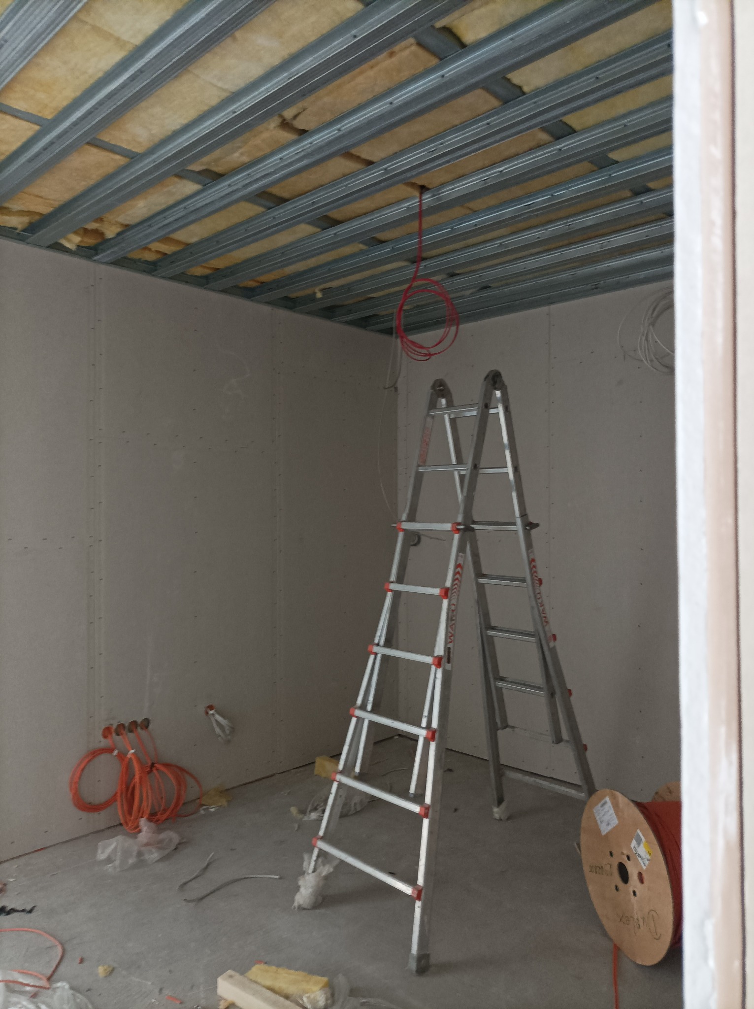 Wnętrze w trakcie prac wykończeniowych: widoczne stelaże sufitowe z izolacją, ściany pokryte płytami gipsowo-kartonowymi, rozłożona drabina aluminiowa, kable elektryczne i zwój kabla na podłodze.