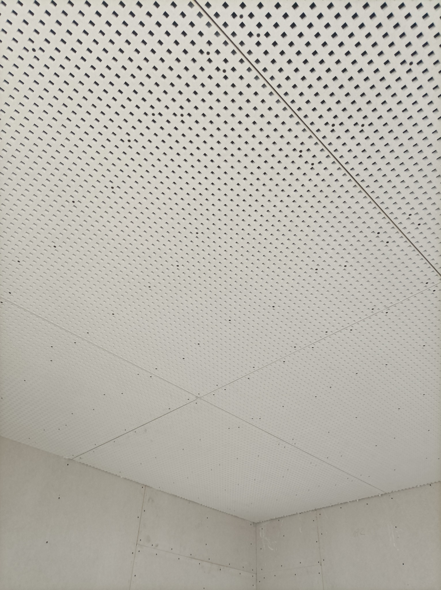 Biały sufit podwieszany z regularnym wzorem kwadratowych perforacji, widoczny w rogu pomieszczenia z surowymi ścianami z płyt kartonowo-gipsowych, z widocznymi śladami po szpachlowaniu i wkrętach.