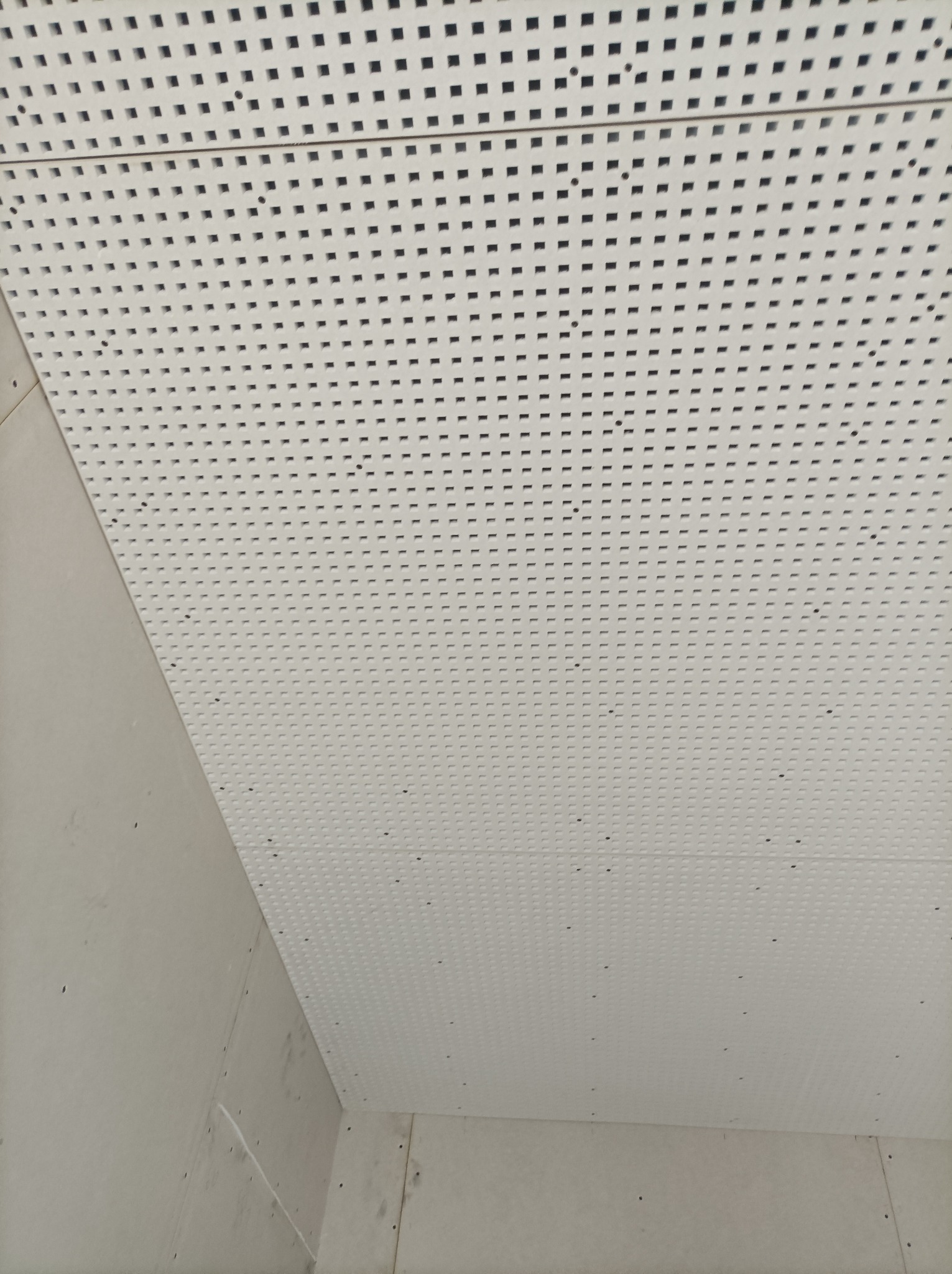 Biały sufit podwieszany z perforacją kwadratową i okrągłą, widoczny z narożnika pomieszczenia z surowymi ścianami.