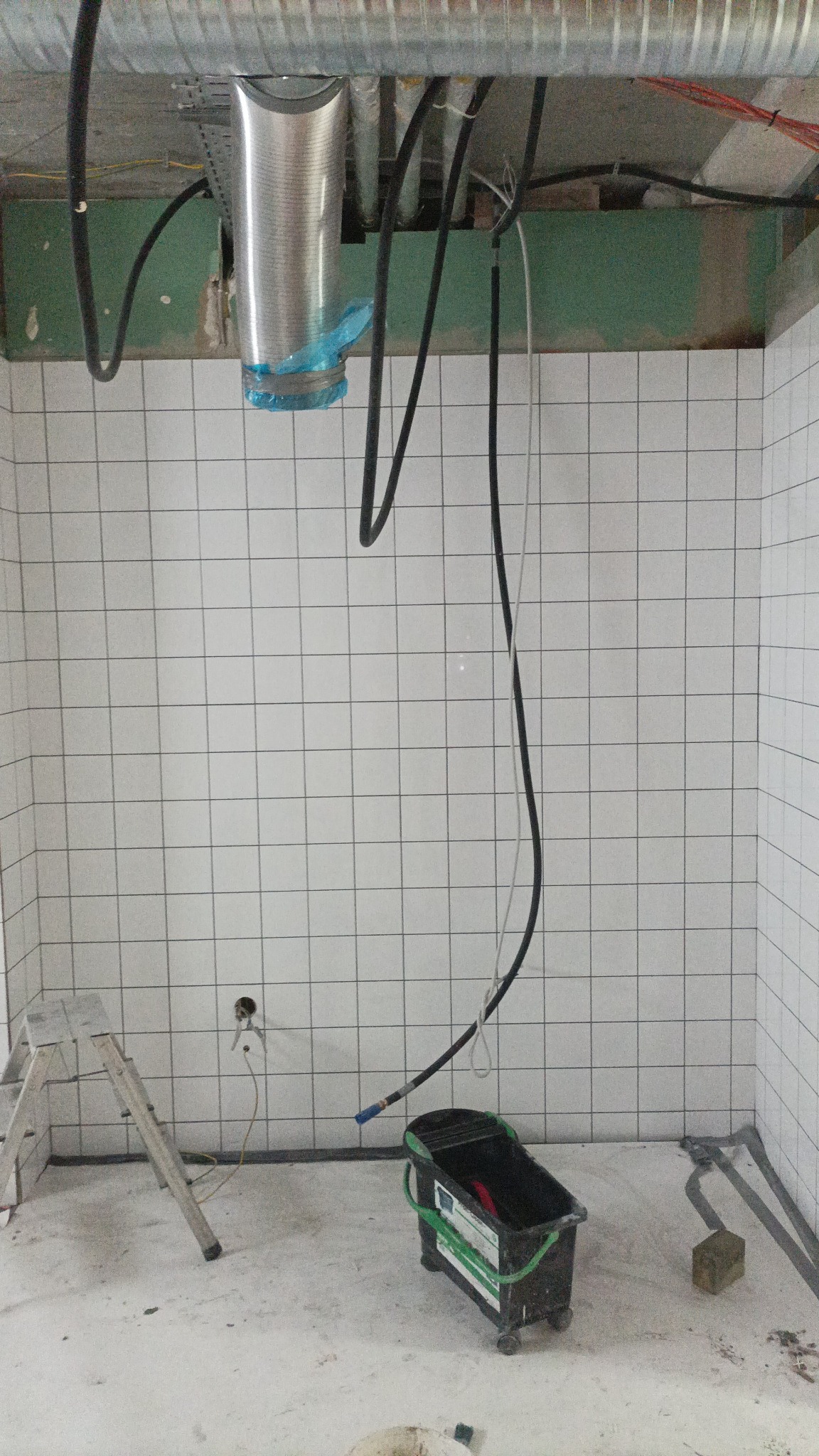 Wnętrze w trakcie prac wykończeniowych: białe płytki na ścianach, aluminiowy kanał wentylacyjny pod sufitem zabezpieczony folią, drabina aluminiowa i wiadro na kółkach z narzędziami na betonowej...