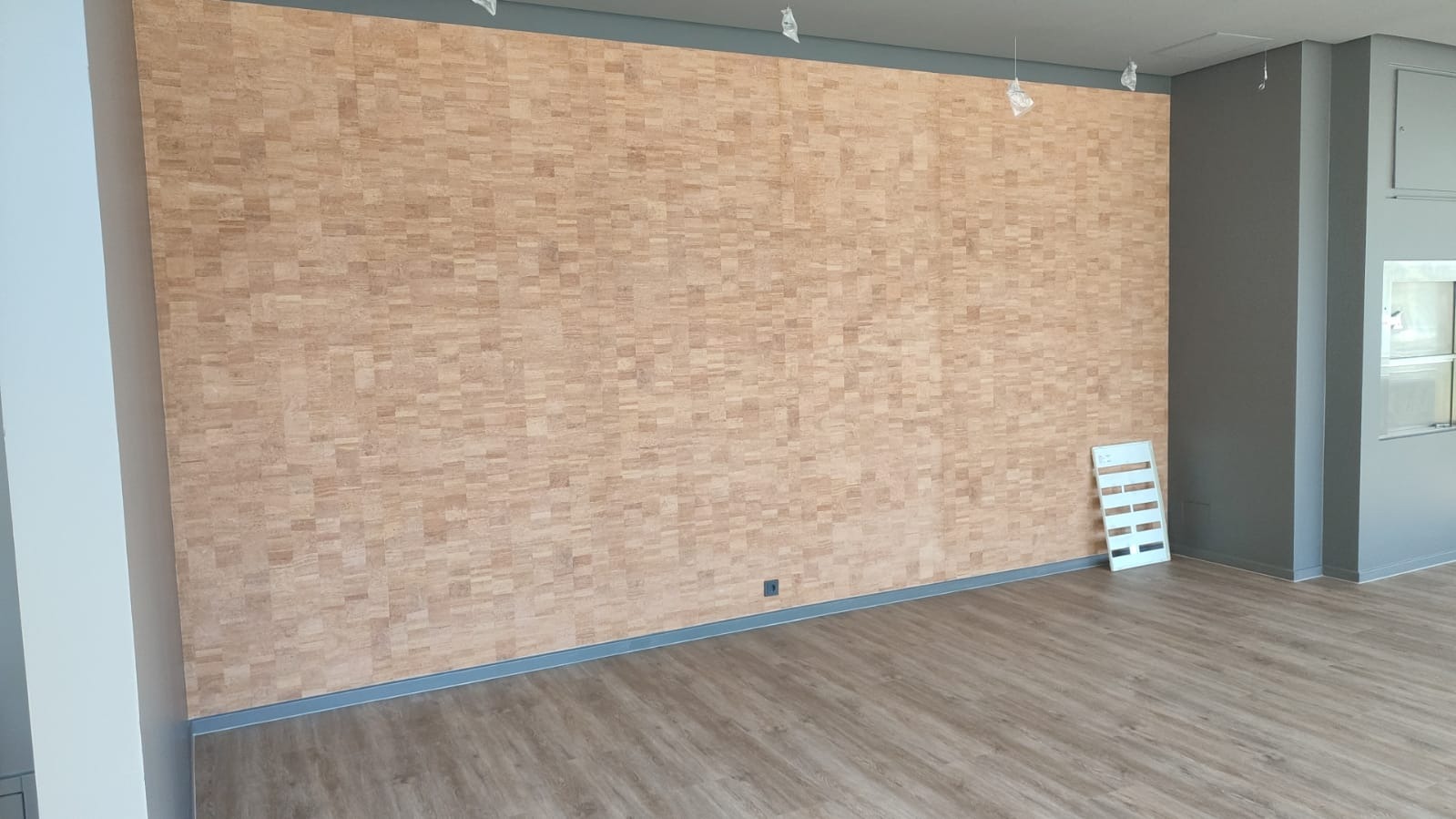 Ściana wykończona korkiem w odcieniach beżu i brązu, z widocznymi drobnymi prostokątnymi elementami, szara listwa przypodłogowa, panele podłogowe imitujące drewno, biała drabinka malarska w rogu...