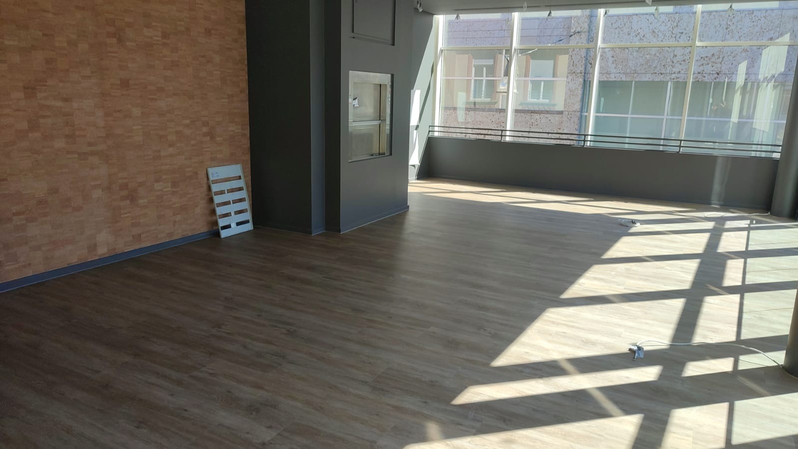 Jasne wnętrze biura z ułożonymi panelami podłogowymi w odcieniach brązu, korkową ścianą i dużymi oknami wpadającymi w geometryczne cienie.