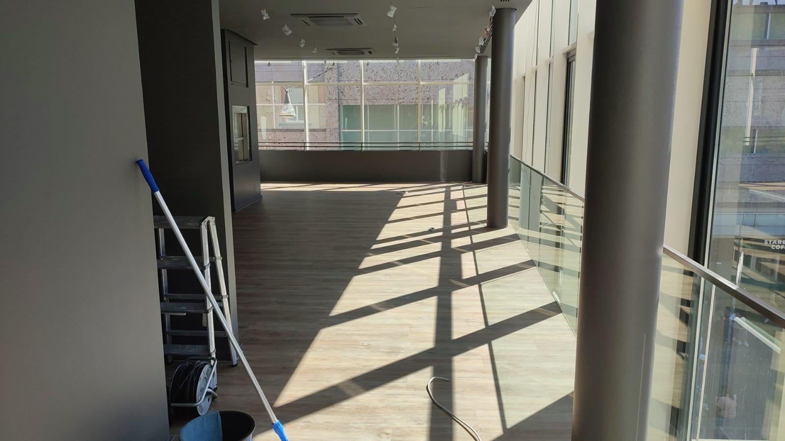 Wnętrze biurowe z panelami podłogowymi w trakcie sprzątania, widoczna drabina, mop i wiadro przy ścianie, duże okna z widokiem na budynek Starbucks.