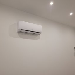 ODNOVA Filip Sidorowicz - Biała jednostka wewnętrzna klimatyzacji marki AUX zamontowana na ścianie w jasnym pomieszczeniu, widoczna skrzynka z bezpiecznikami w prawym dolnym rogu.