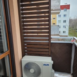 ODNOVA Filip Sidorowicz - Jednostka zewnętrzna klimatyzacji AUX zamontowana na balkonie w Jaczowie, osłonięta drewnianą przesłoną, z widokiem na blok mieszkalny w tle.
