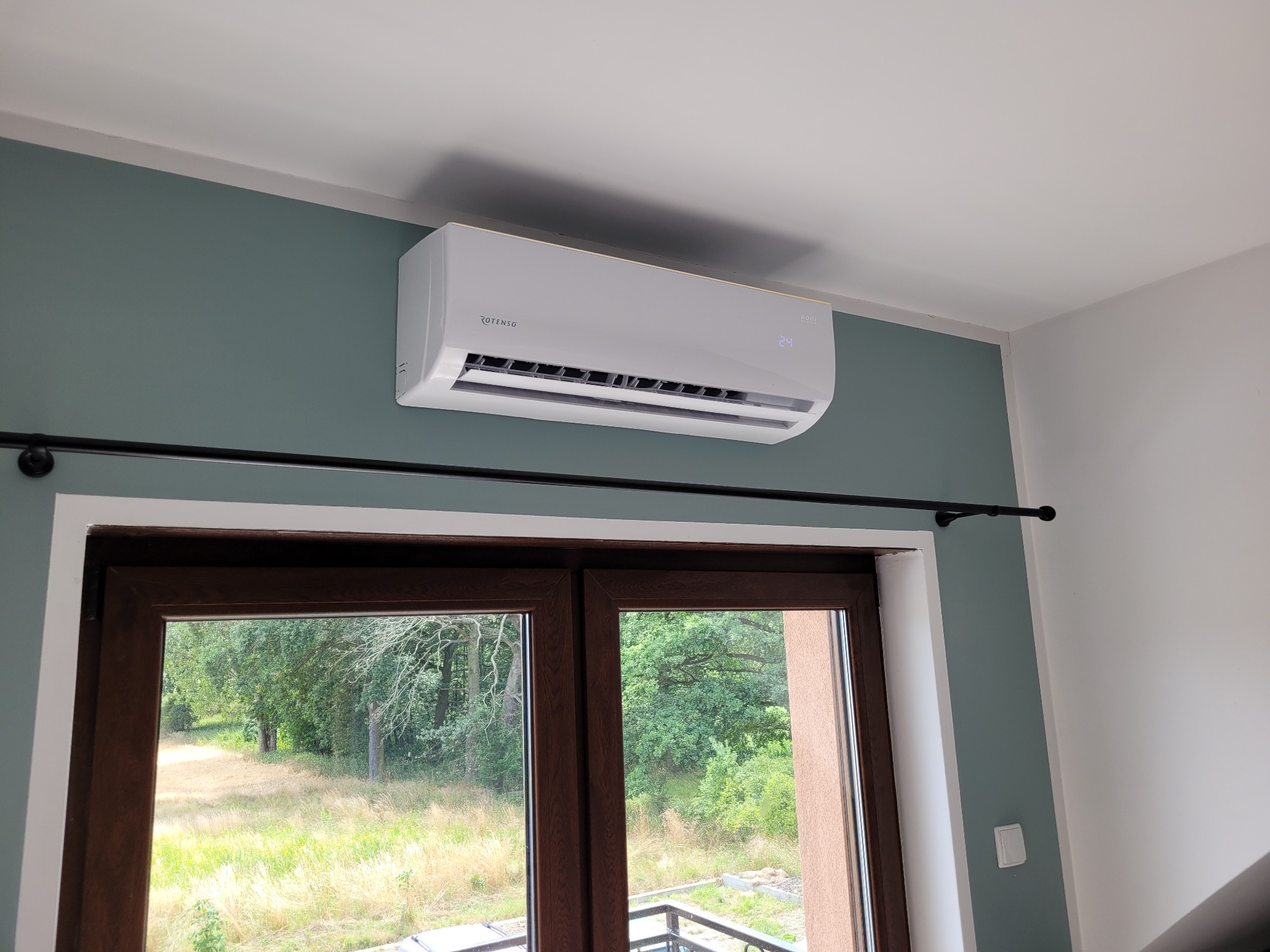 Biały klimatyzator marki Rotenso zamontowany na zielonej ścianie w pokoju z widokiem na zewnątrz przez brązowe okno.