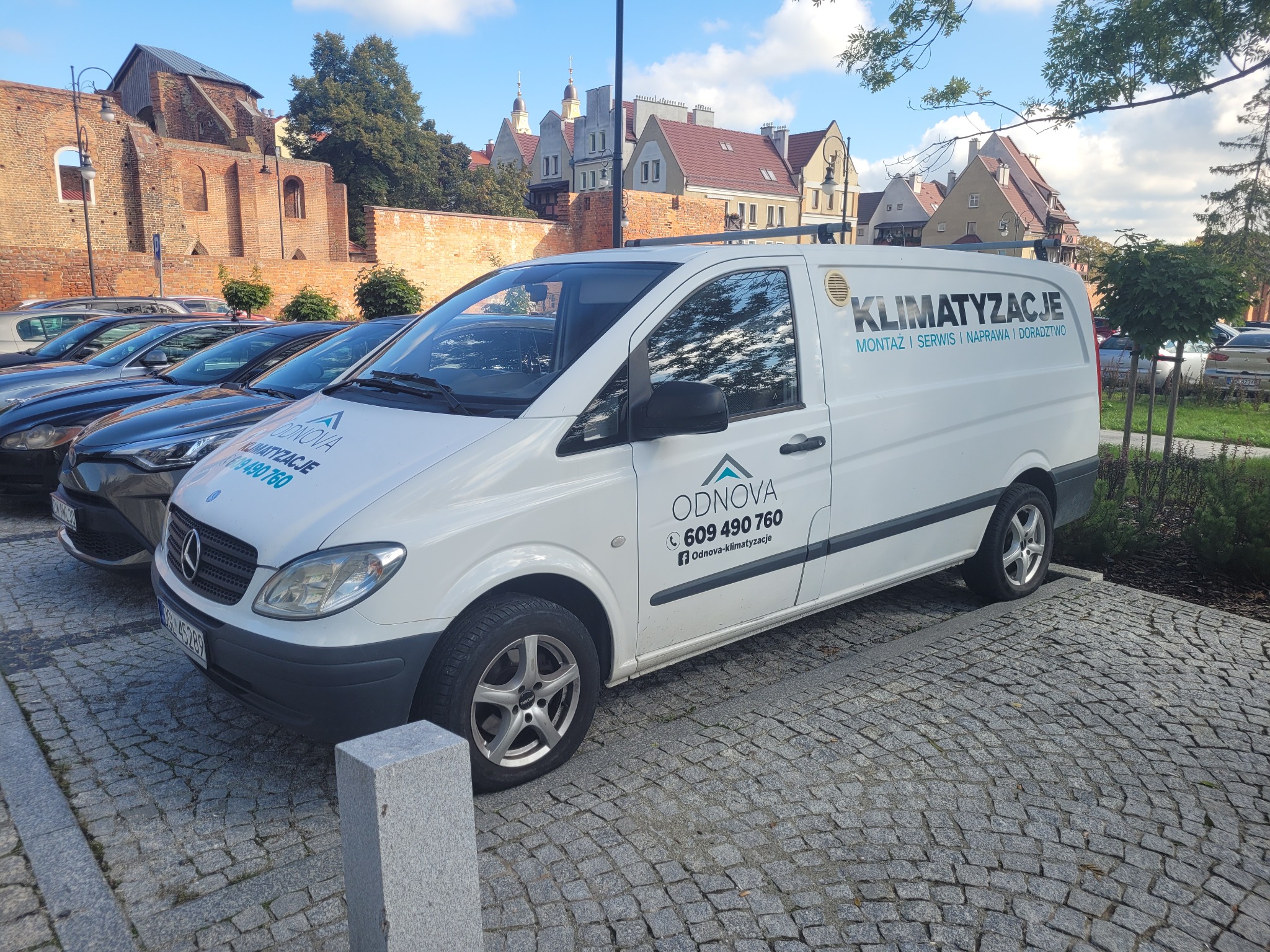 Biały samochód dostawczy firmy świadczącej usługi montażu i serwisu klimatyzacji, zaparkowany na brukowanej ulicy w tle zabytkowa zabudowa miejska.