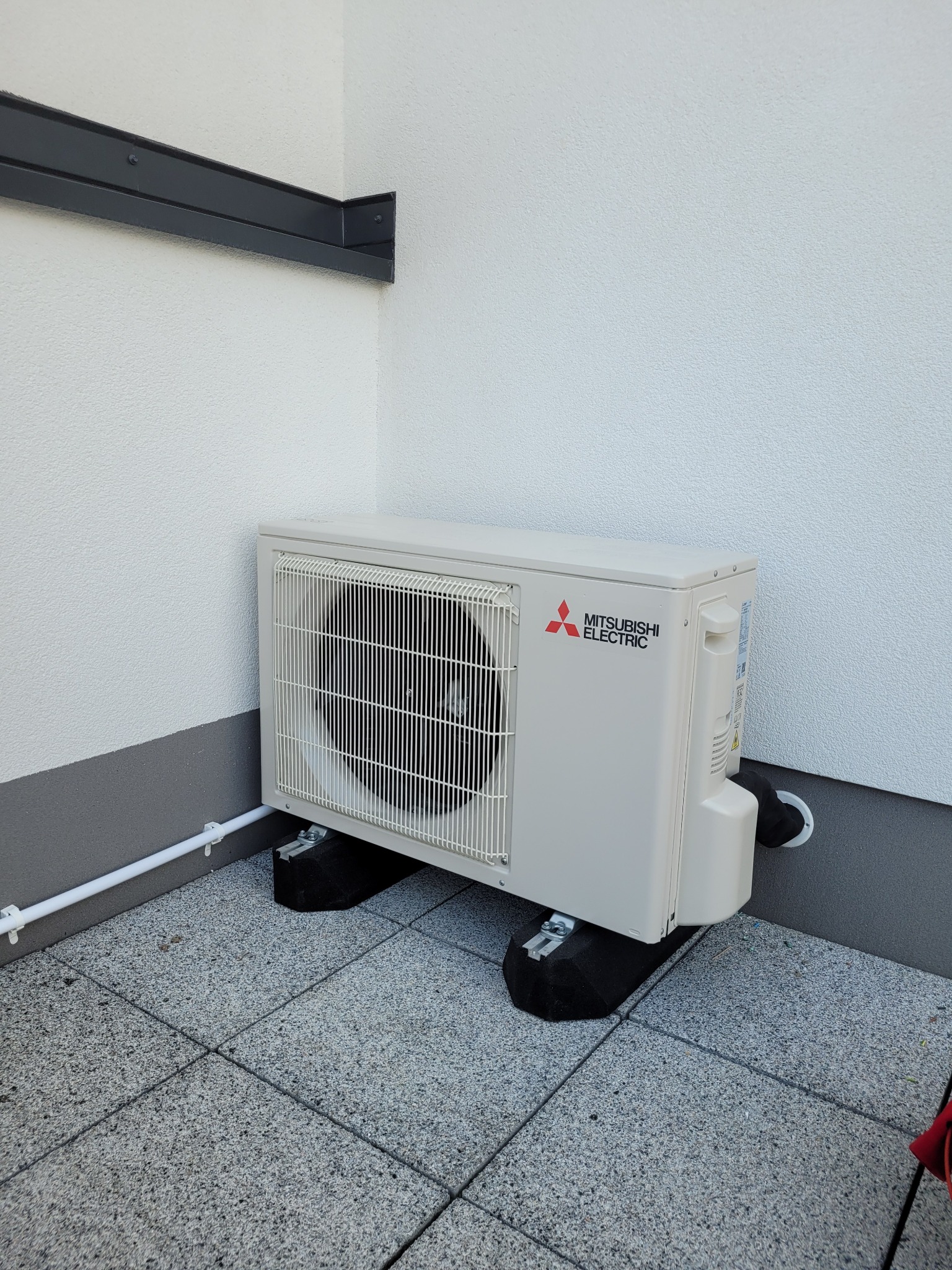 Zewnętrzna jednostka klimatyzacji Mitsubishi Electric zamontowana na grafitowych podkładkach antywibracyjnych, z widoczną białą rurą odprowadzającą skropliny, na tle jasnej, teksturowanej ściany...