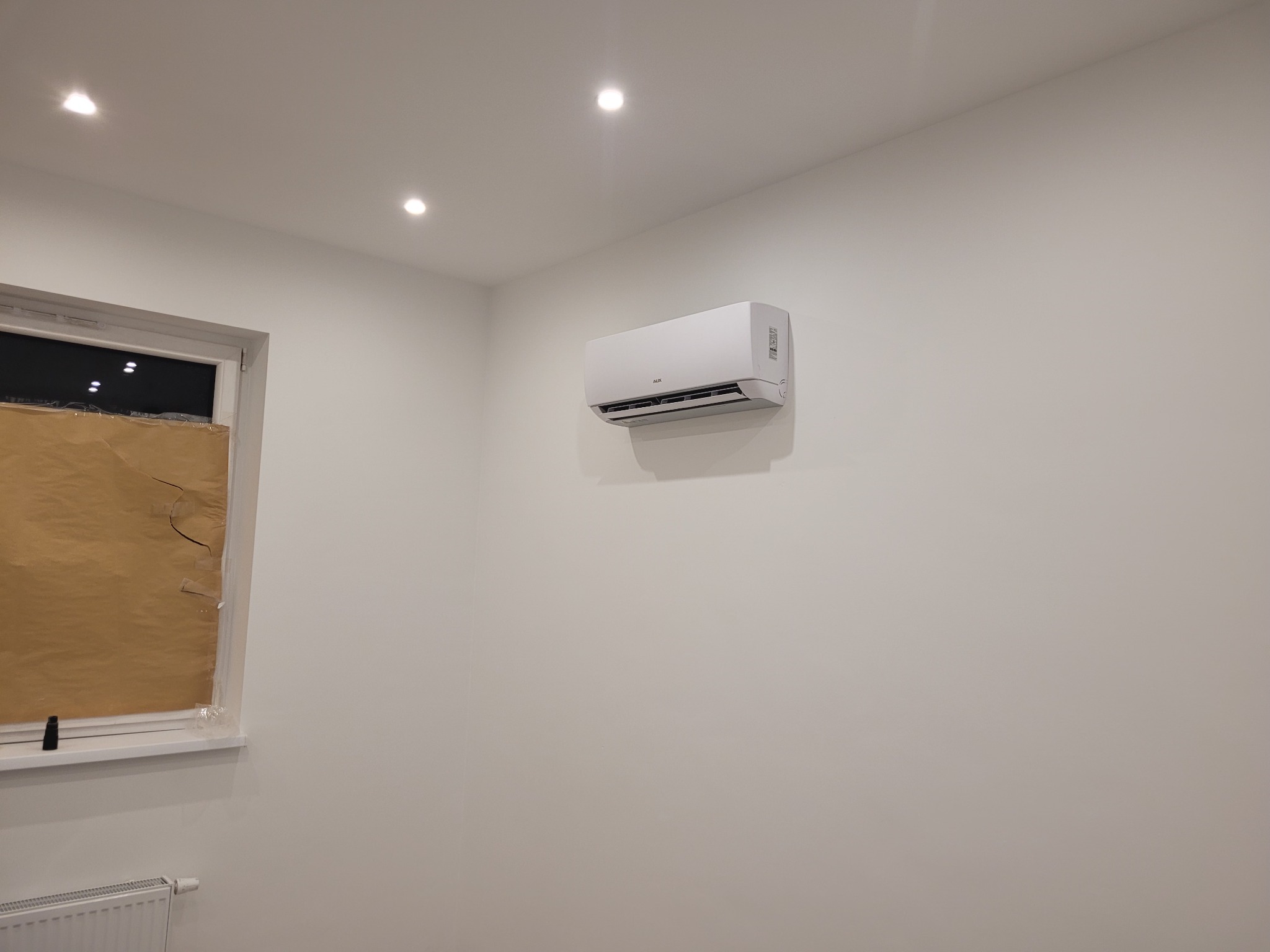 Biały klimatyzator ścienny marki AUX zamontowany w jasnym pomieszczeniu z oknem zasłoniętym brązowym papierem ochronnym.