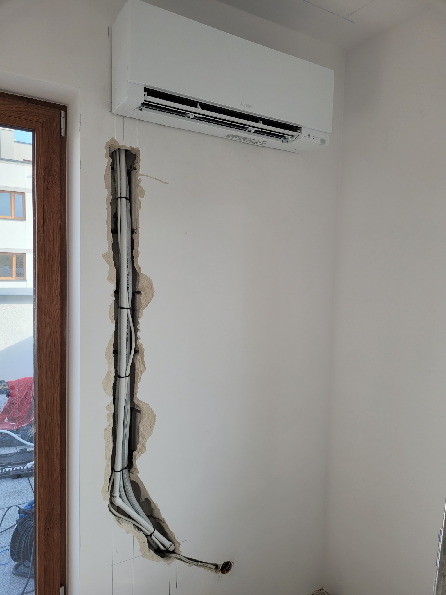 Biała jednostka wewnętrzna klimatyzacji zamontowana na ścianie w nowym budynku. Widoczna bruzda w ścianie z przewodami instalacyjnymi, przygotowana do zakrycia. Świeżo otynkowana ściana.
