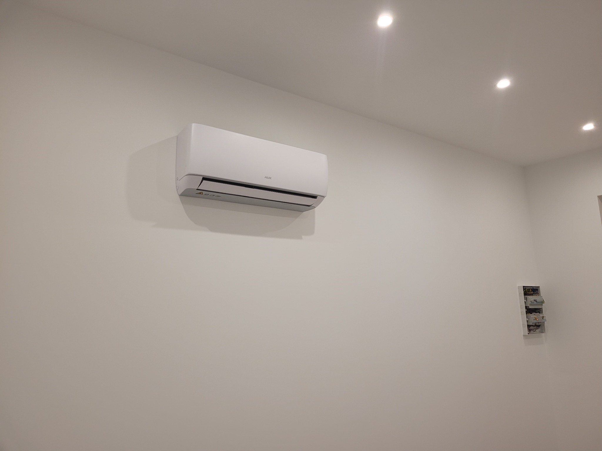 Biała jednostka wewnętrzna klimatyzacji marki AUX zamontowana na ścianie w jasnym pomieszczeniu, widoczna skrzynka z bezpiecznikami w prawym dolnym rogu.