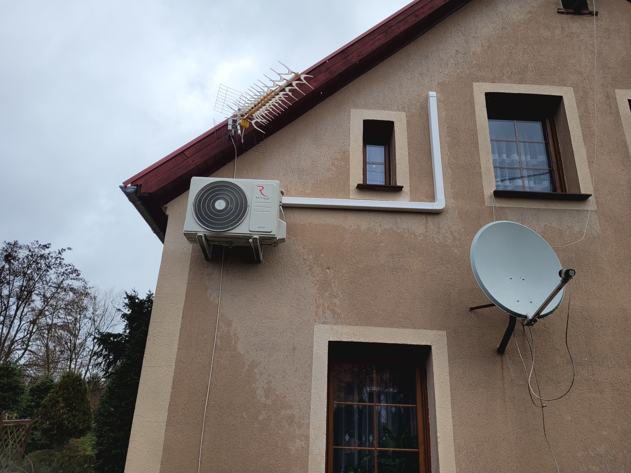Zewnętrzna jednostka klimatyzacji Rotenso zamontowana na elewacji domu jednorodzinnego, widoczna antena telewizyjna i satelitarna.