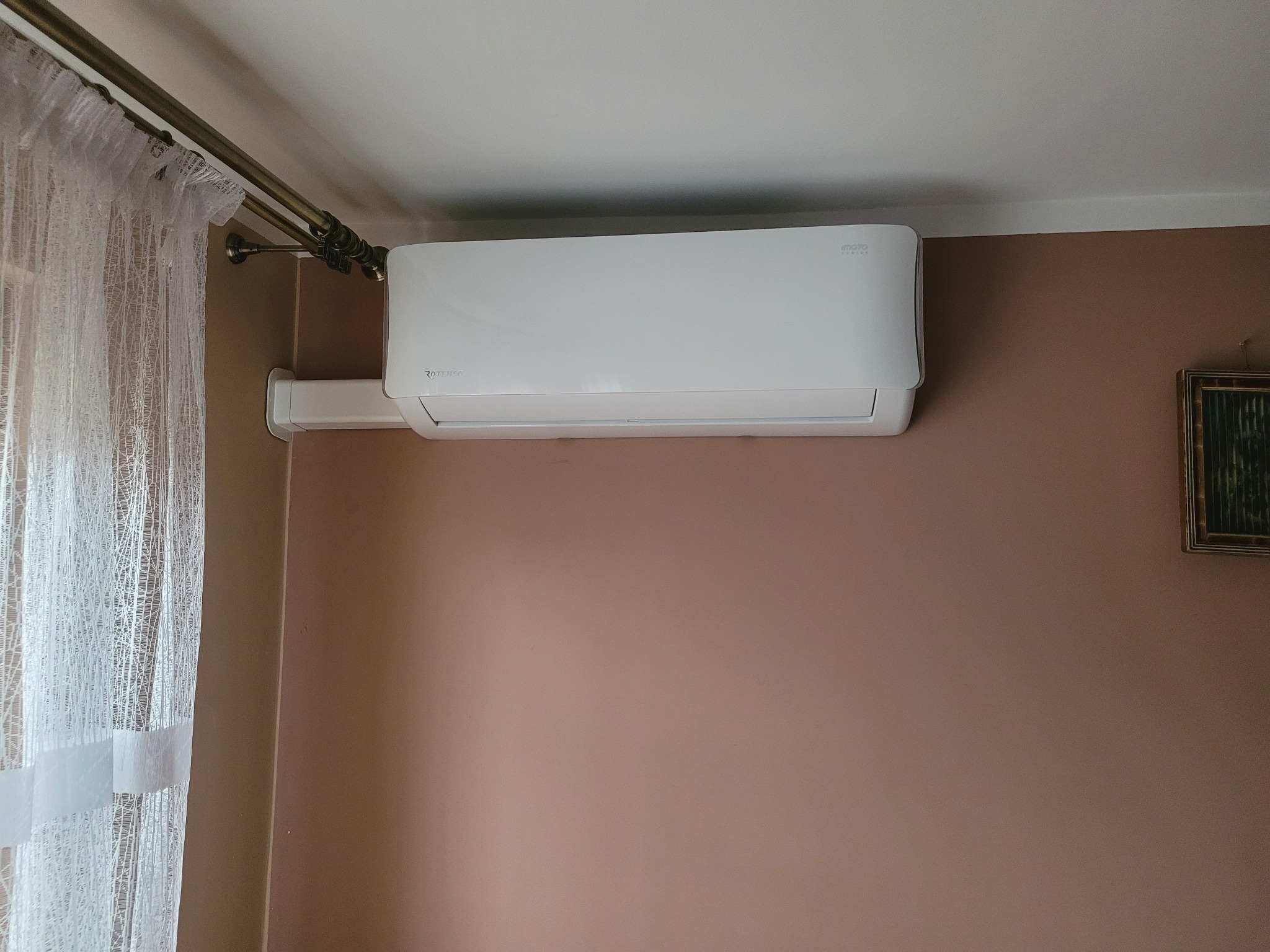 Biały klimatyzator Rotenso zamontowany na brązowej ścianie w pokoju, widoczne firanki i obrazek w ramce.