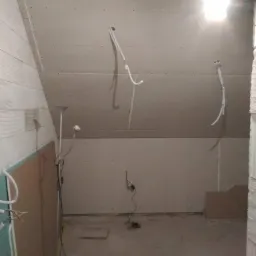 Poddasze w trakcie wykańczania płytami kartonowo-gipsowymi, widoczne przewody elektryczne wystające ze ścian i sufitu, ściana z białego bloczka oraz podłoga w stanie surowym.