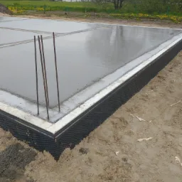 Świeżo wylana, mokra płyta fundamentowa z wystającymi prętami zbrojeniowymi, zabezpieczona izolacją pionową z folii kubełkowej na obrzeżach, otoczona ziemią.