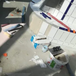 Prace w toku: układanie białych i niebieskich płytek w zaokrąglonym basenie, widoczne narzędzia, poziomica, puszki z zaprawą, resztki materiałów i akcesoriów na podłodze.