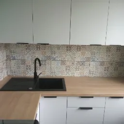 Kuchnia po renowacji: białe szafki z czarnymi uchwytami, drewniany blat i mozaika z płytek nad zlewem z czarną armaturą.