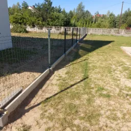 Nowoczesne ogrodzenie panelowe z podmurówką betonową, widok z trawnika w słoneczny dzień, w tle drzewa i dom.