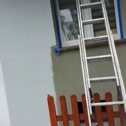 Częściowo pomalowana elewacja budynku w odcieniach szarości, zabezpieczone okno taśmą malarską, drabina oparta o ścianę, widoczny fragment drewnianego płotu i karton na ziemi.