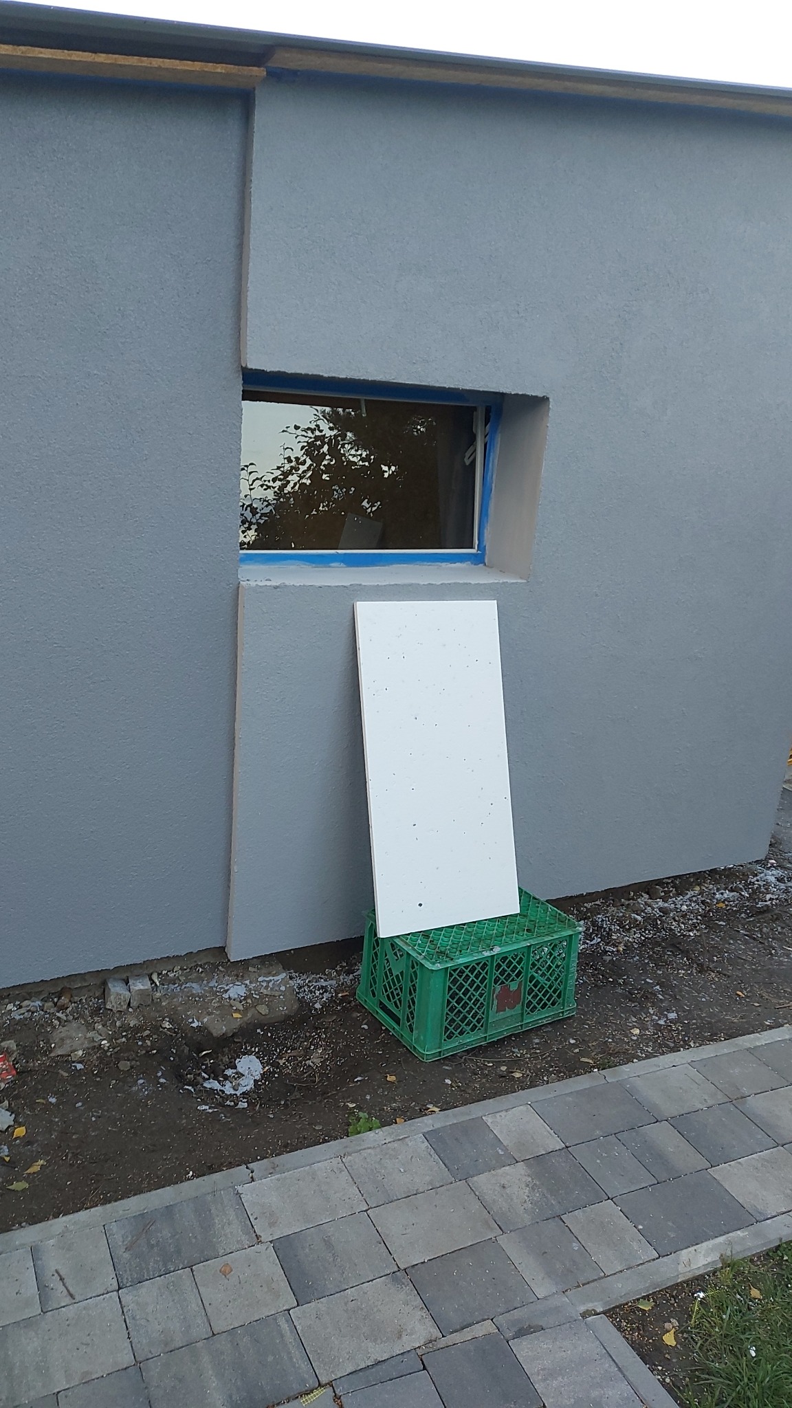 Świeżo pomalowana szara ściana zewnętrzna z oknem zabezpieczonym taśmą malarską; biała płyta oparta o ścianę na zielonej skrzynce.