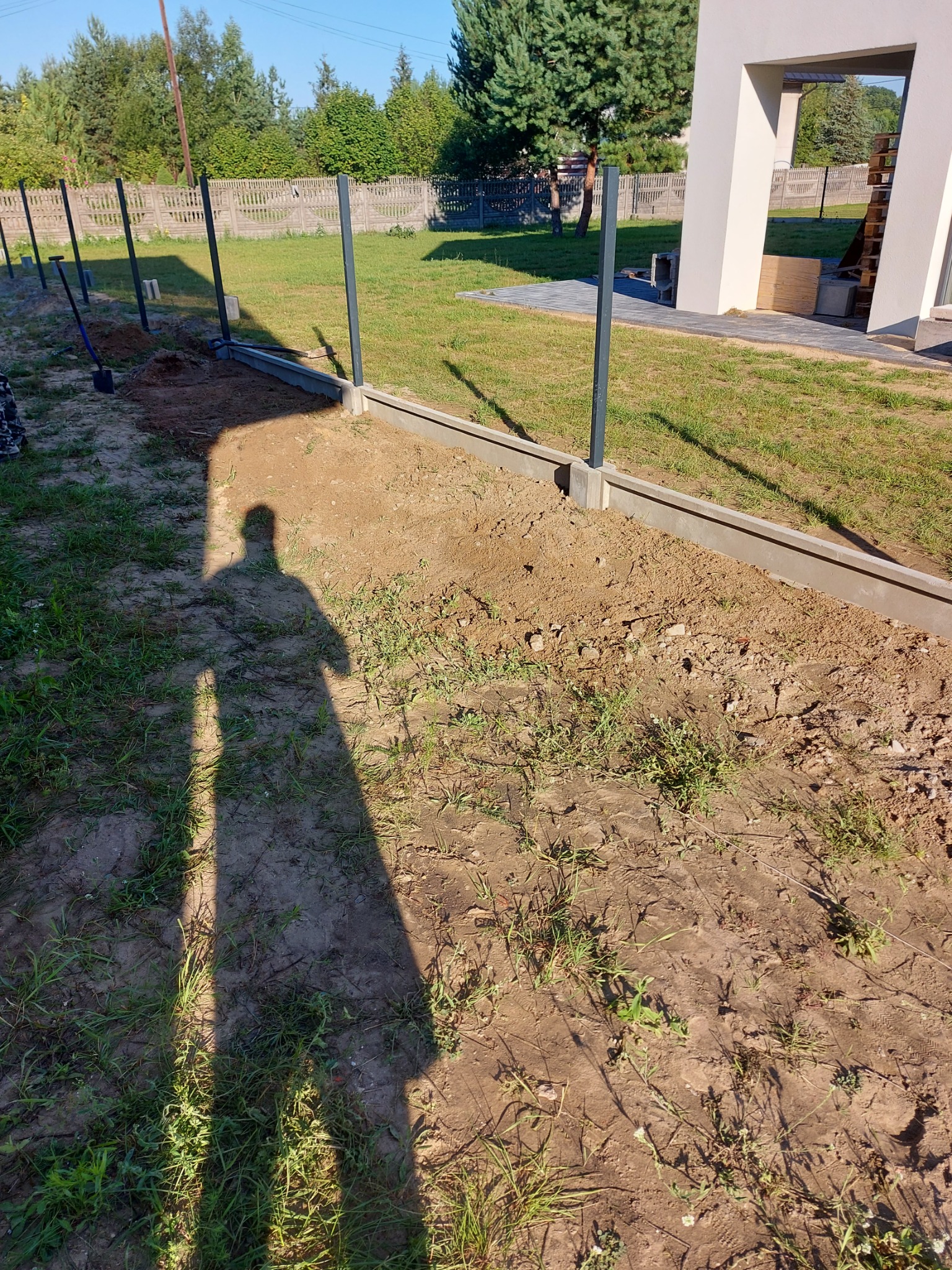 Montaż słupków ogrodzeniowych na betonowej podmurówce, widoczny cień osoby wykonującej prace, teren częściowo przygotowany pod ogrodzenie.
