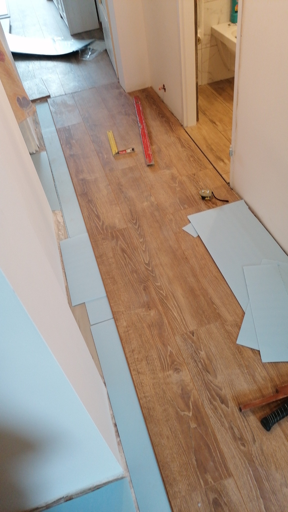 Układanie paneli podłogowych w korytarzu, widoczne narzędzia pomiarowe i fragment łazienki z umywalką w tle.