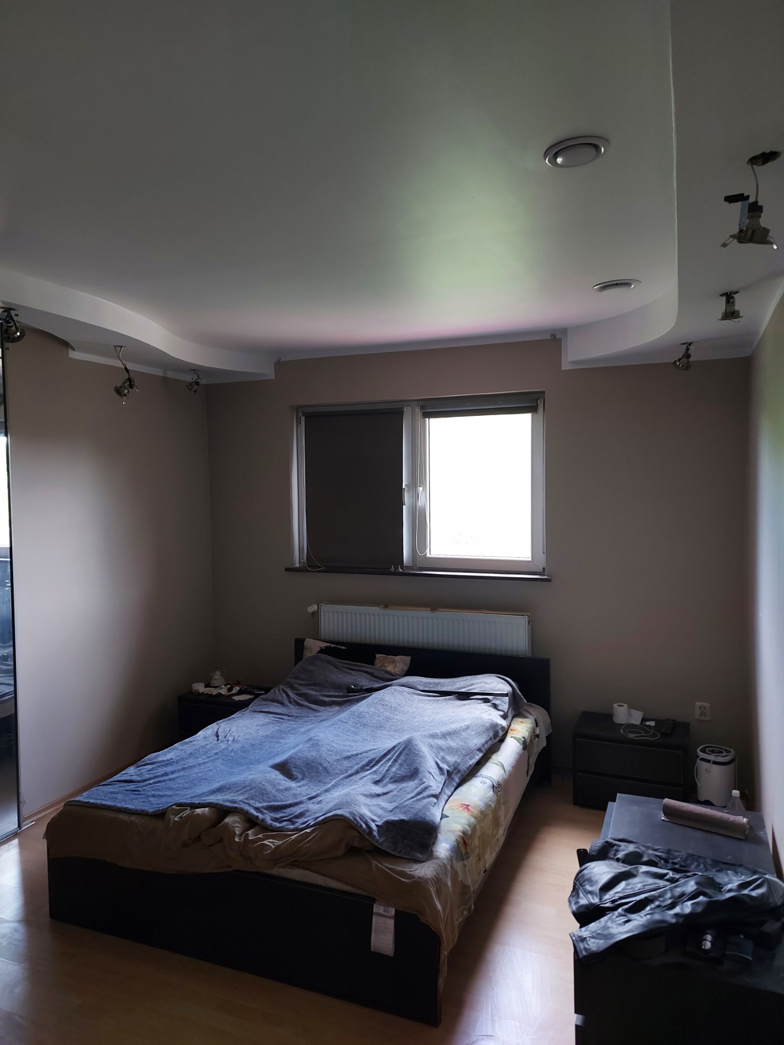 Pokój z beżowymi ścianami, łóżkiem z niebieską narzutą i podwieszanym sufitem z oświetleniem punktowym, okno z roletą.