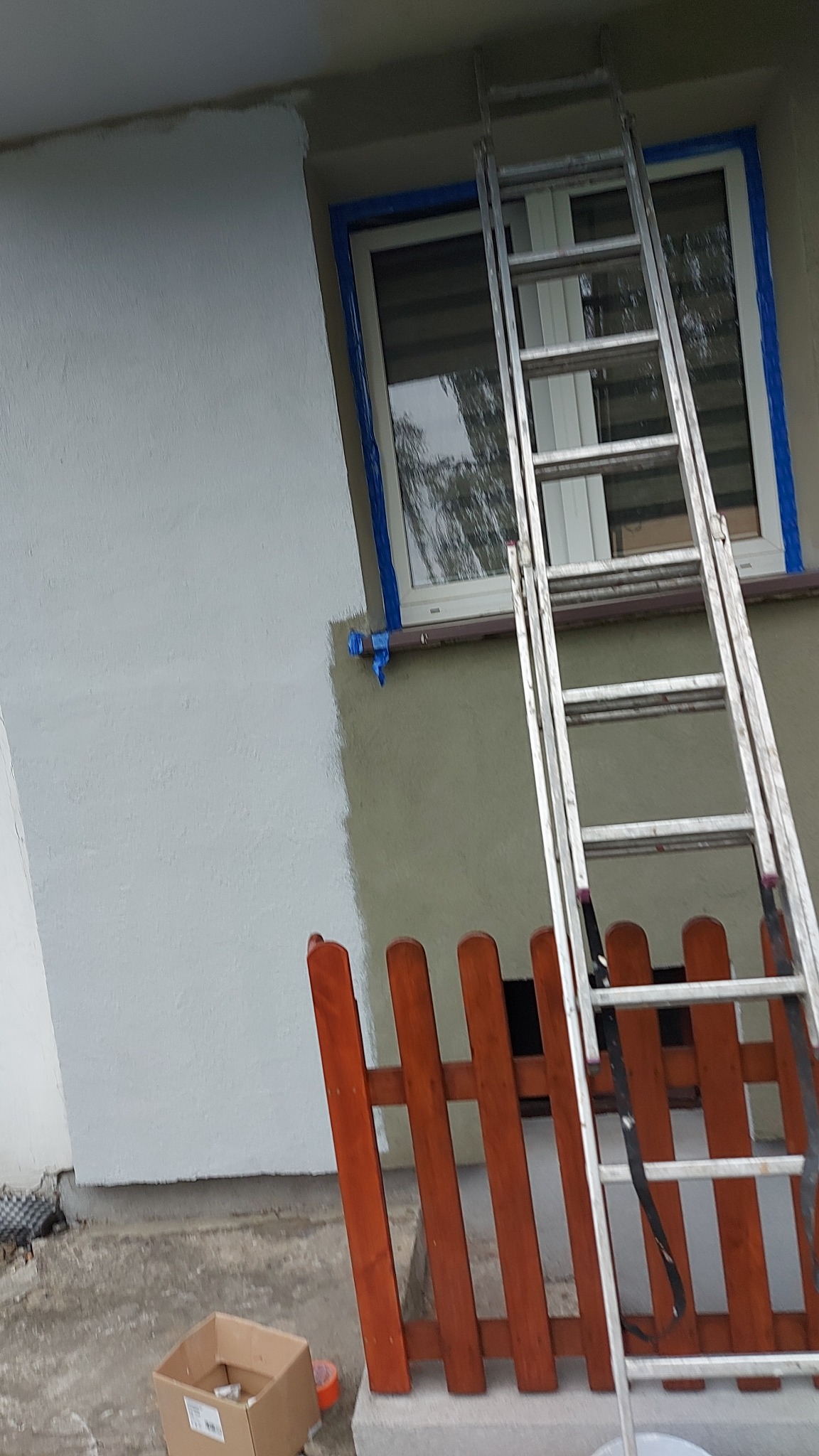 Częściowo pomalowana elewacja budynku w odcieniach szarości, zabezpieczone okno taśmą malarską, drabina oparta o ścianę, widoczny fragment drewnianego płotu i karton na ziemi.