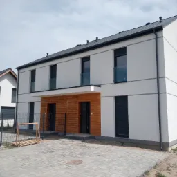 Nowoczesny, dwukondygnacyjny dom z białymi ścianami, drewnianą elewacją wejściową i szarymi roletami zewnętrznymi, widok z zewnątrz na kostkę brukową i częściowo zagospodarowany teren.