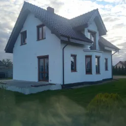 Dwukondygnacyjny dom z białymi ścianami i ciemnobrązowym dachem, widoczny z boku na tle pochmurnego nieba.