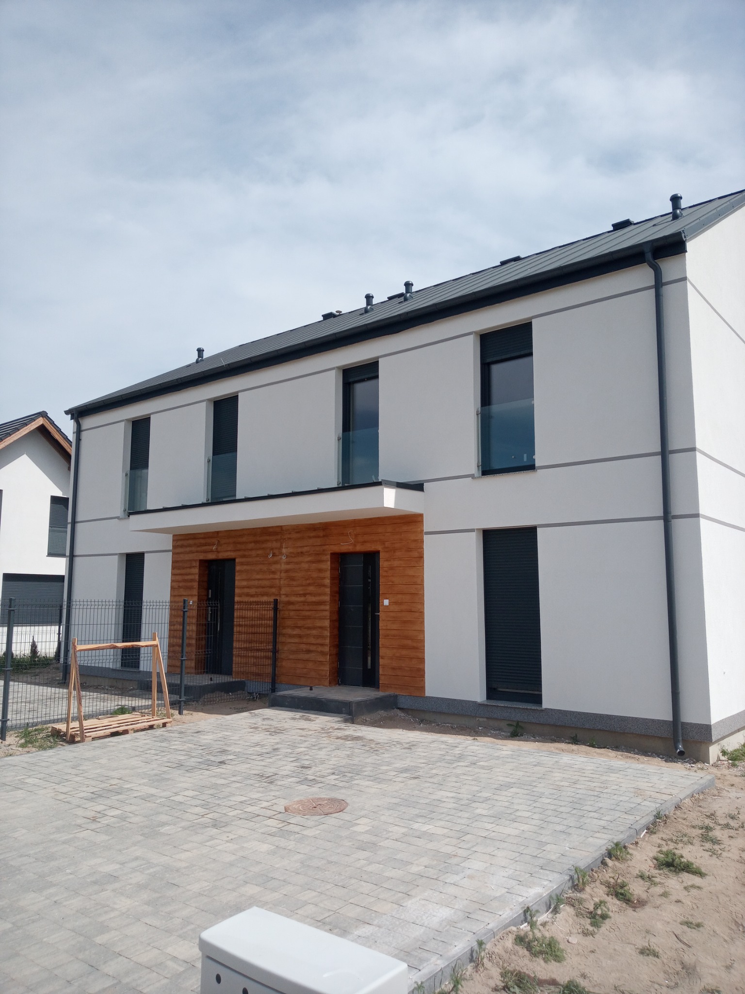 Nowoczesny, dwukondygnacyjny dom z białymi ścianami, drewnianą elewacją wejściową i szarymi roletami zewnętrznymi, widok z zewnątrz na kostkę brukową i częściowo zagospodarowany teren.
