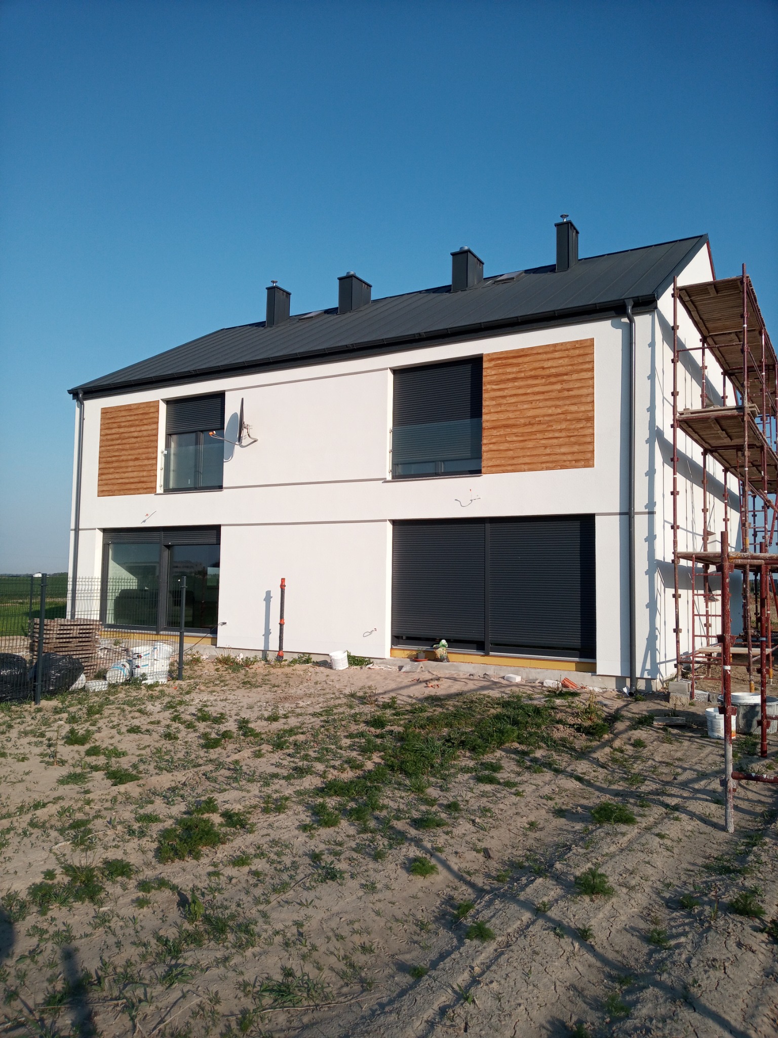 Dwukondygnacyjny dom z białymi ścianami, drewnianymi panelami dekoracyjnymi i szarą stolarką okienną, częściowo zasłoniętą roletami zewnętrznymi, widziany z zewnątrz na tle błękitnego nieba,...