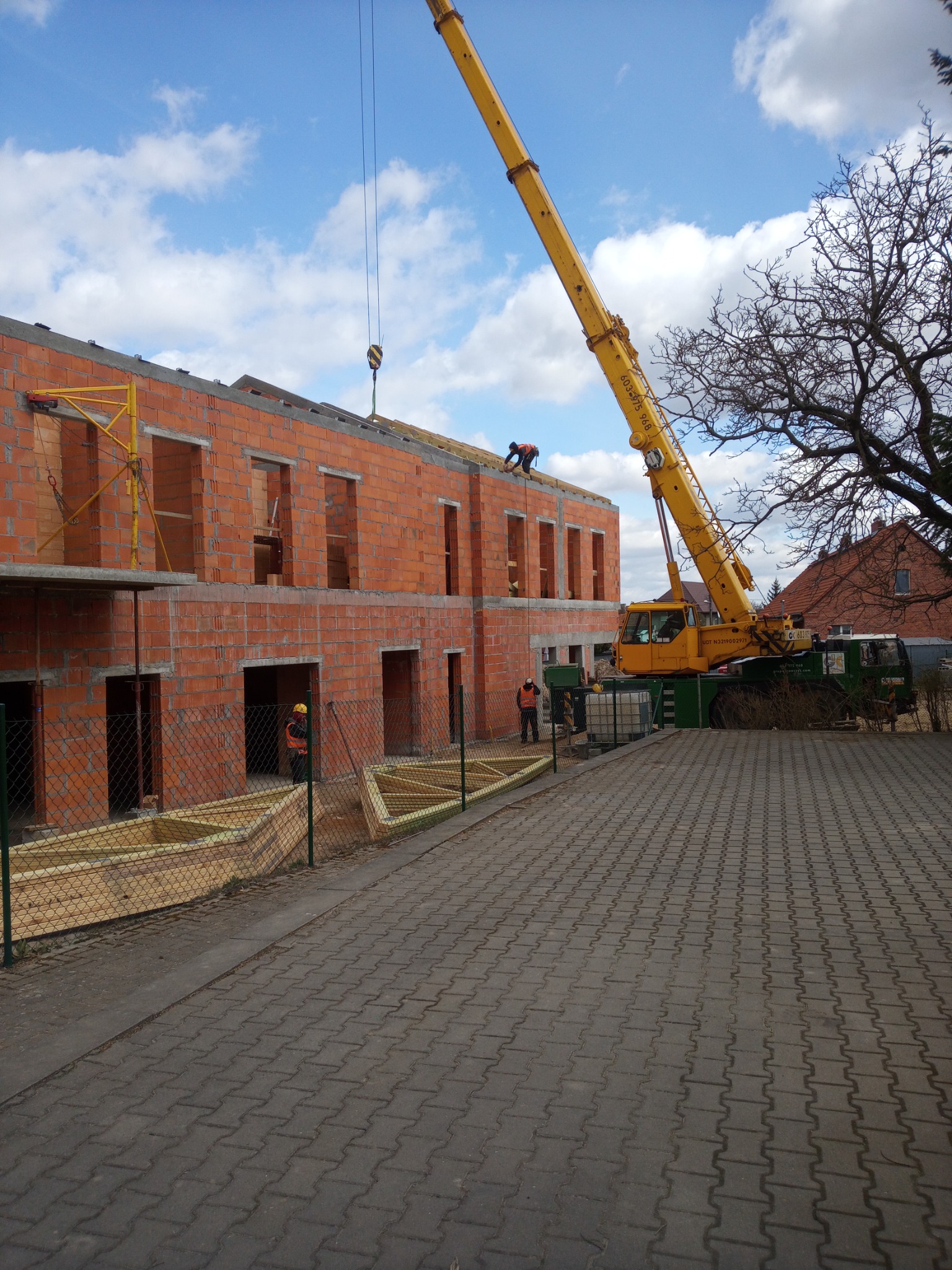 Budowa domu z czerwonej cegły, widoczny dźwig podający elementy konstrukcyjne na dach, pracownicy w kamizelkach odblaskowych i kaskach, budynek w stanie surowym otwartym.