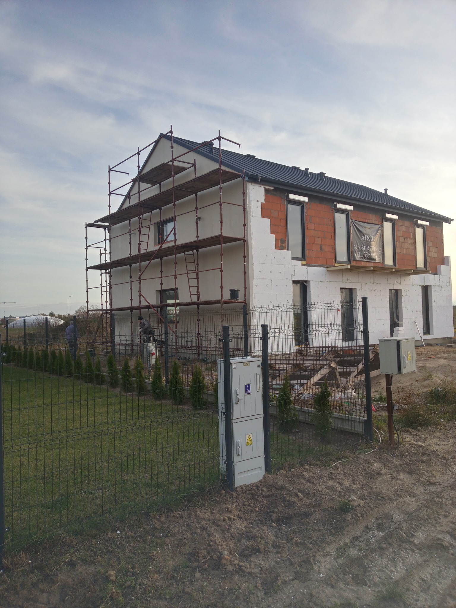 Dom w trakcie ocieplania: widoczne rusztowania, częściowo ocieplona elewacja z białym styropianem, fragment ściany z czerwonej cegły, okna z czarnymi ramami, baner reklamowy na elewacji, pracownicy...