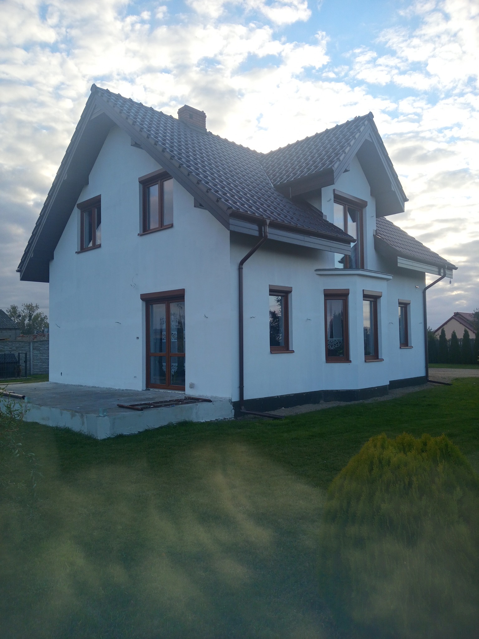 Dwukondygnacyjny dom z białymi ścianami i ciemnobrązowym dachem, widoczny z boku na tle pochmurnego nieba.