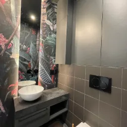 Nowoczesna łazienka z umywalką nablatową i szafką pod umywalką w kolorze grafitowym, ściana z motywem tropikalnym i grafitowe szafki wiszące, widoczny przycisk spłuczki toalety.