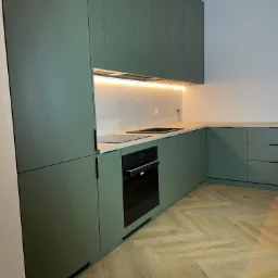 Nowoczesna kuchnia z zielonymi szafkami, drewnianym blatem i parkietem w jodełkę, oświetlenie LED pod szafkami górnymi.