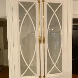 Biała, wysoka szafa z przeszklonymi drzwiami z ozdobnymi, złotymi uchwytami. Na szufladach widoczne taśmy malarskie.