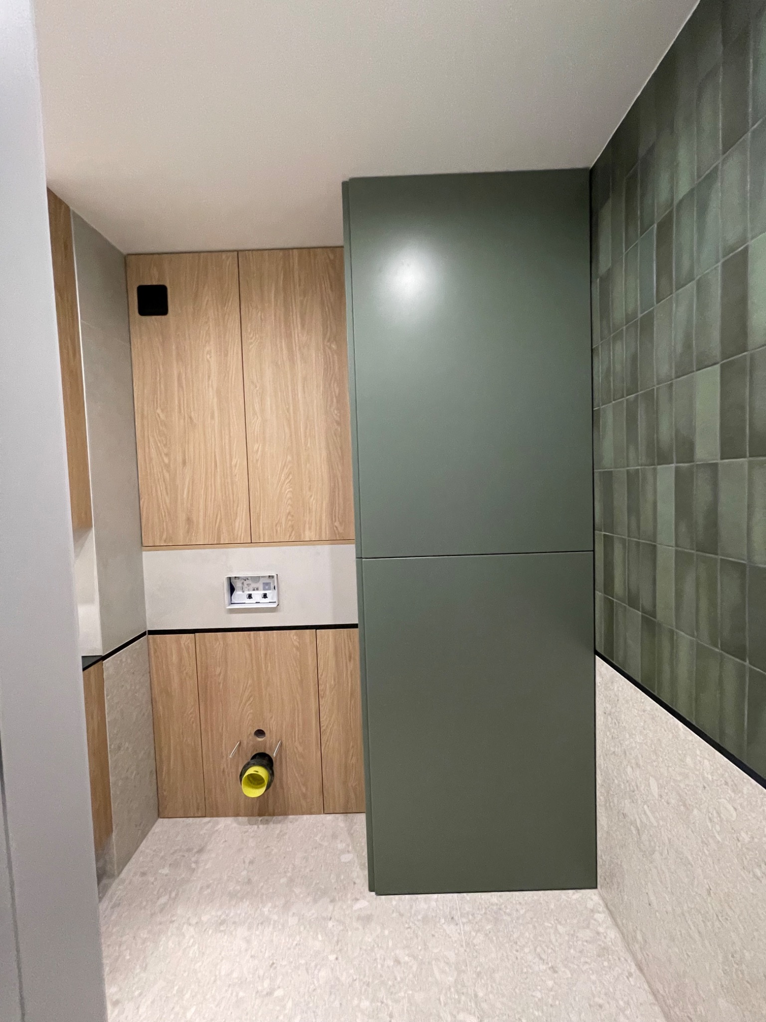 Nowoczesna łazienka w trakcie wykańczania: drewnopodobne szafki, zielona zabudowa, płytki w odcieniach zieleni, widoczne instalacje sanitarne.