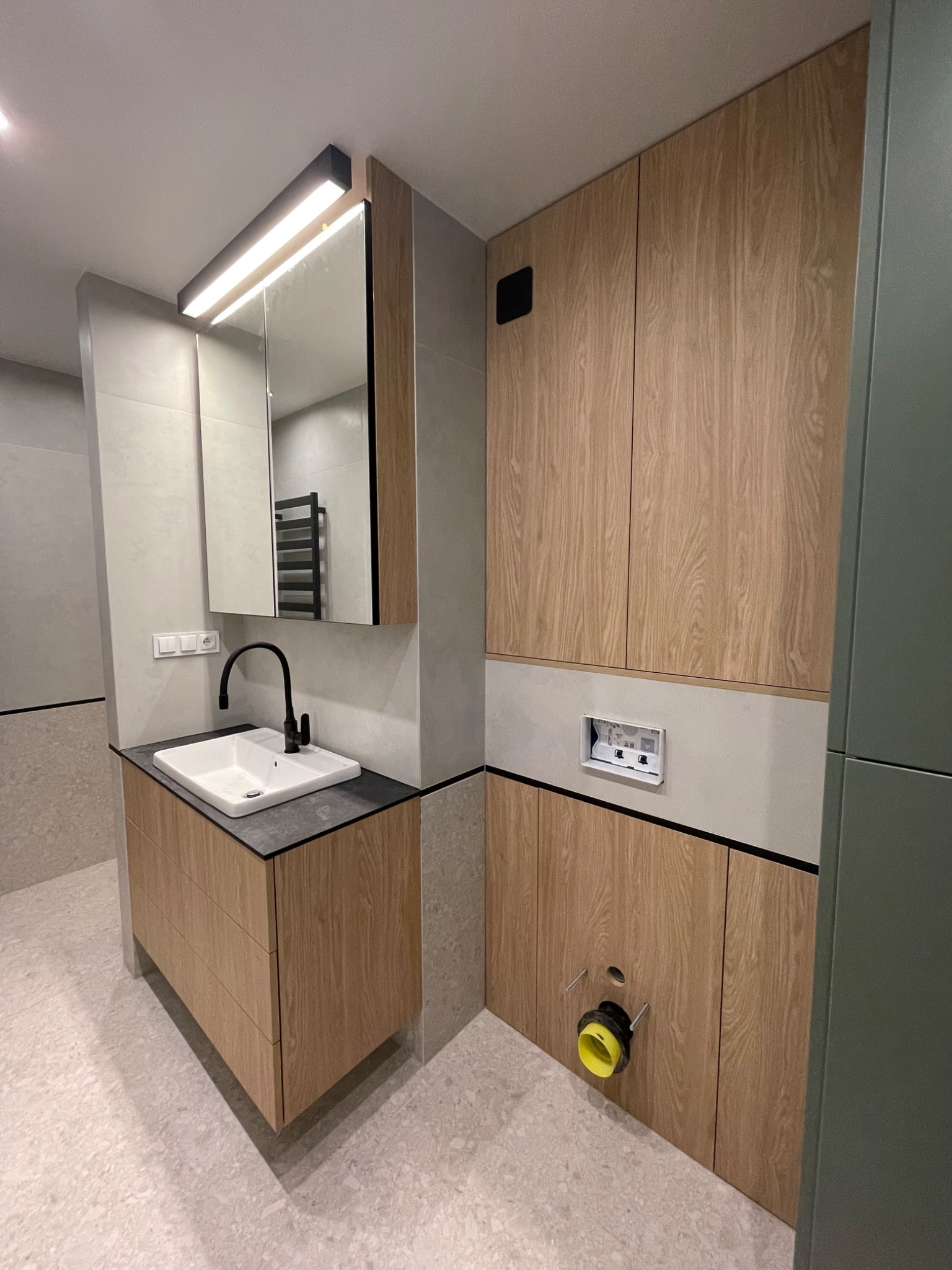 Nowoczesna łazienka z drewnianymi szafkami na wymiar, czarną armaturą i podświetlanym lustrem, z widocznym odpływem kanalizacyjnym z żółtym elementem.