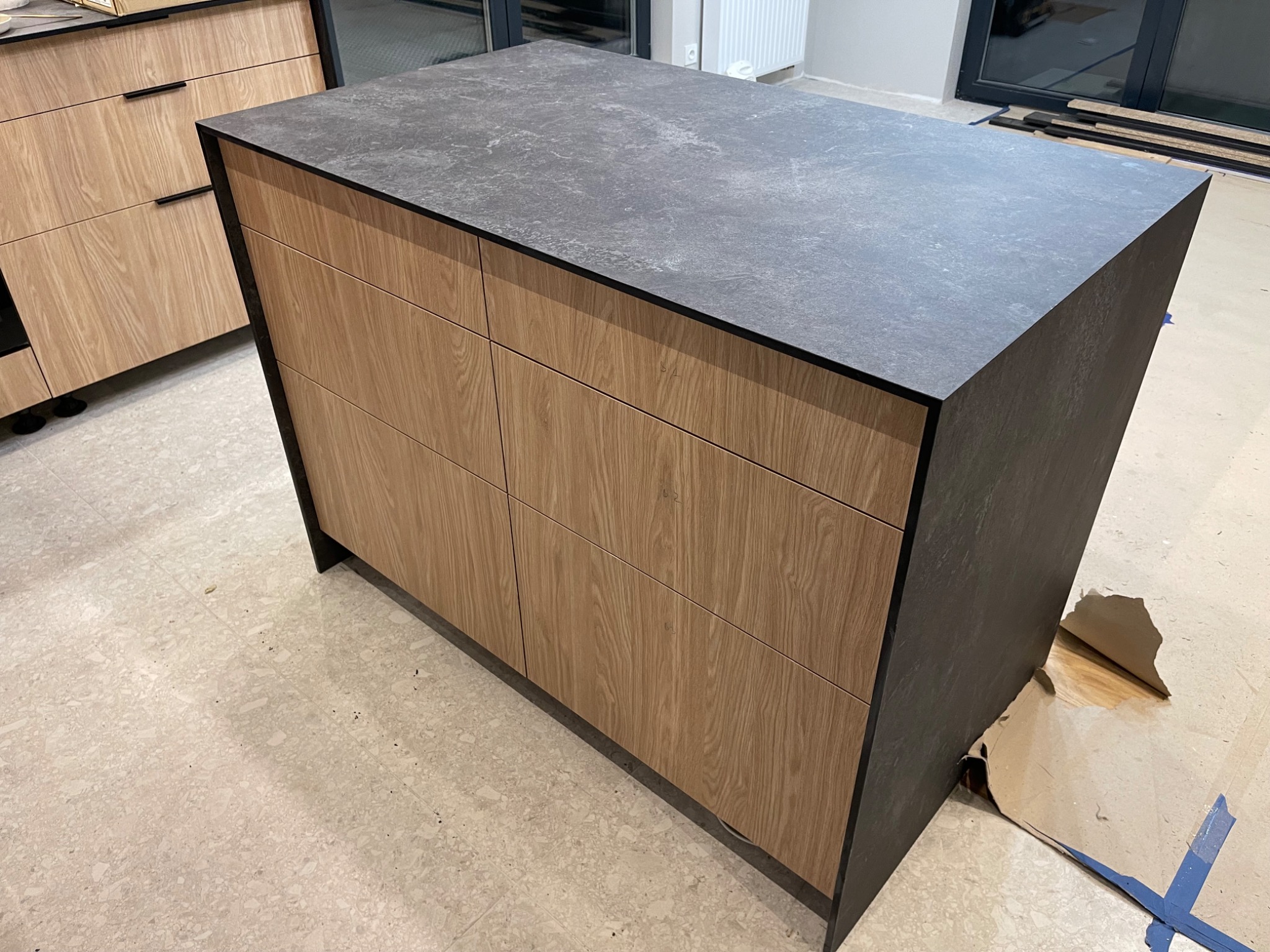 Wyspa kuchenna z drewnopodobnymi frontami szuflad i ciemnoszarym blatem, widoczne kartony zabezpieczające podłogę podczas montażu.