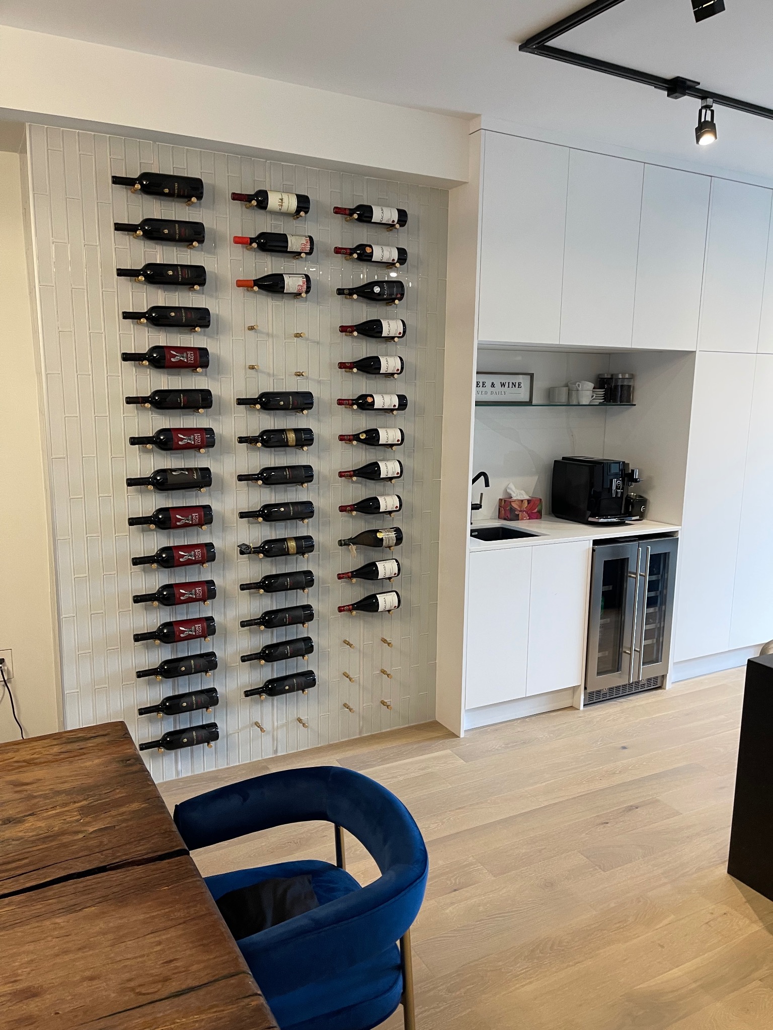Ściana z białych płytek z rzędami butelek wina na złotych uchwytach, obok biała zabudowa kuchenna z barkiem na wino i ekspresem do kawy, drewniany stół i niebieskie krzesło.