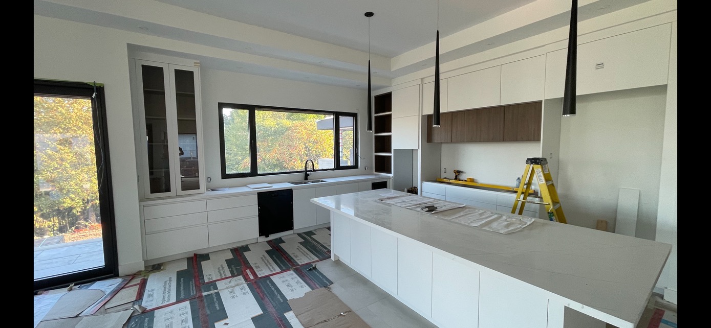Wnętrze nowoczesnej kuchni w trakcie montażu, z białą zabudową i wyspą, czarną armaturą i oknem z widokiem na drzewa jesienią, zabezpieczona podłoga, widoczna drabina i narzędzia.