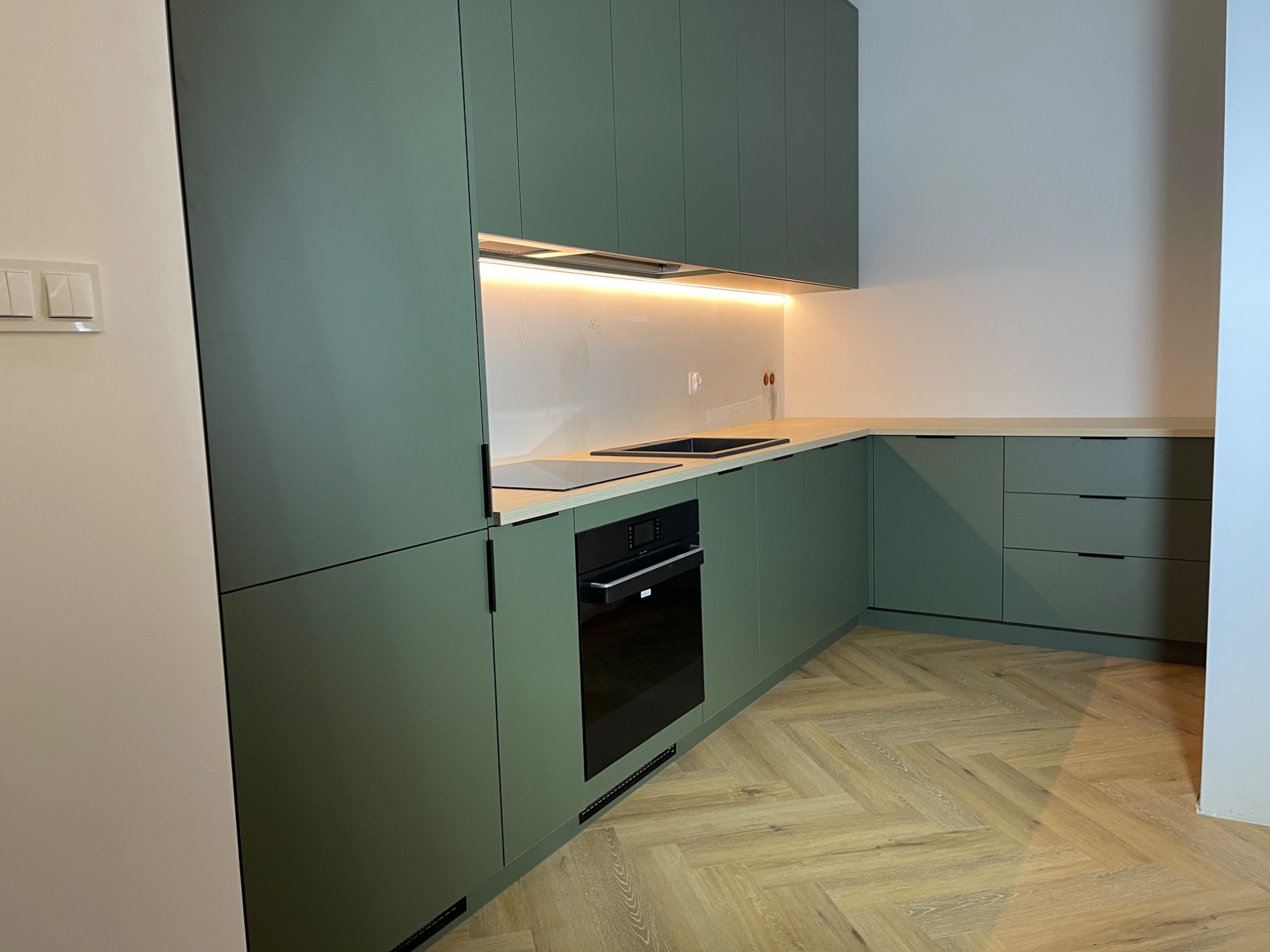 Nowoczesna kuchnia z zielonymi szafkami, drewnianym blatem i parkietem w jodełkę, oświetlenie LED pod szafkami górnymi.