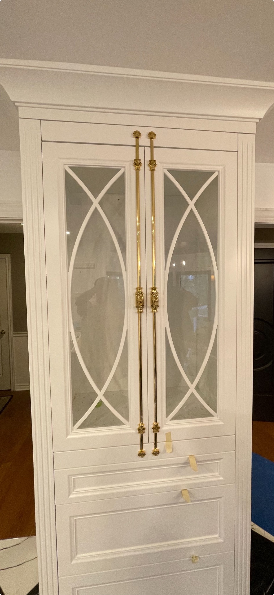 Biała, wysoka szafa z przeszklonymi drzwiami z ozdobnymi, złotymi uchwytami. Na szufladach widoczne taśmy malarskie.