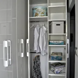 Biała szafa wnękowa z połyskiem, otwarte drzwi ukazują półki z dekoracyjnymi pudełkami, wiszące białe koszule i sukienkę w kwiaty, na dole para błyszczących szpilek.